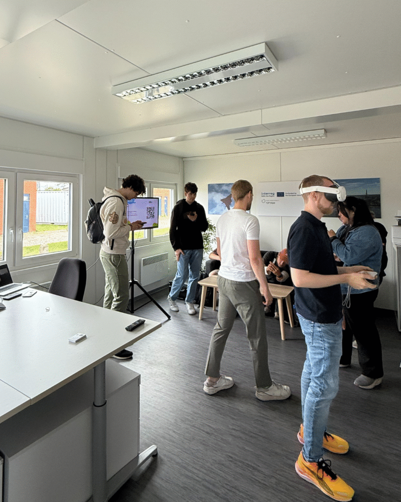 People gathered in a modern meeting room with large windows; some standing and using smartphones or VR headsets, while others sit near a table. A screen shows a slide with a QR code, and EU project posters are on the wall.