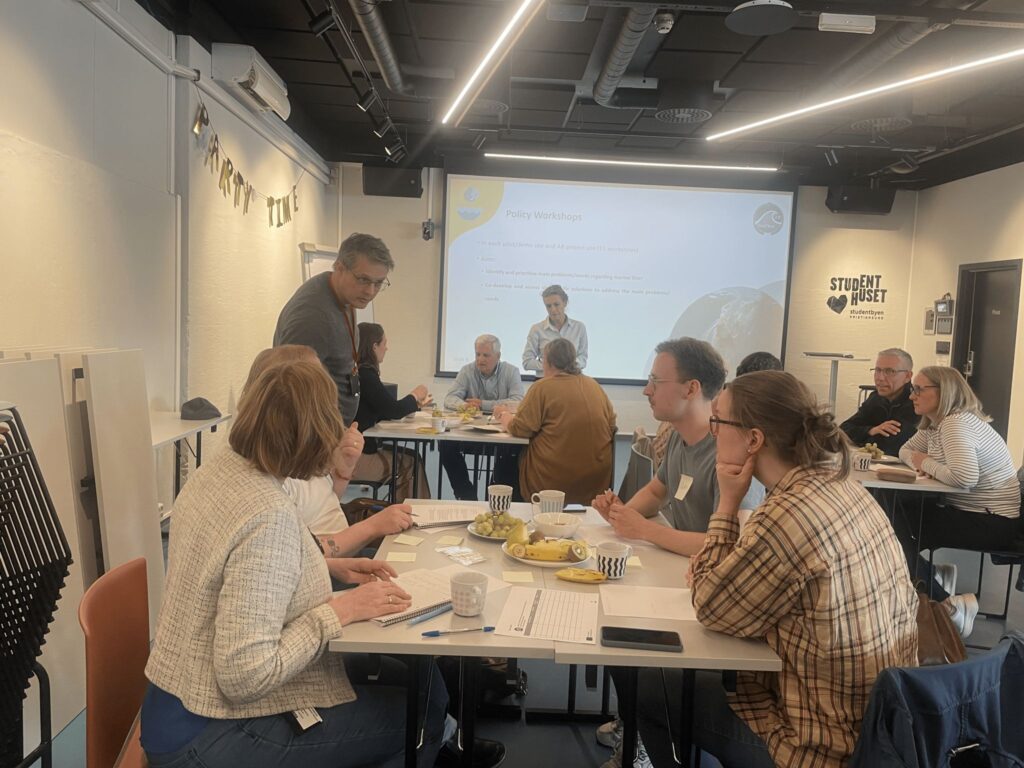 Participants sit in groups around tables during a policy workshop, discussing and taking notes, while a presentation is projected on a large screen at the front of the room.