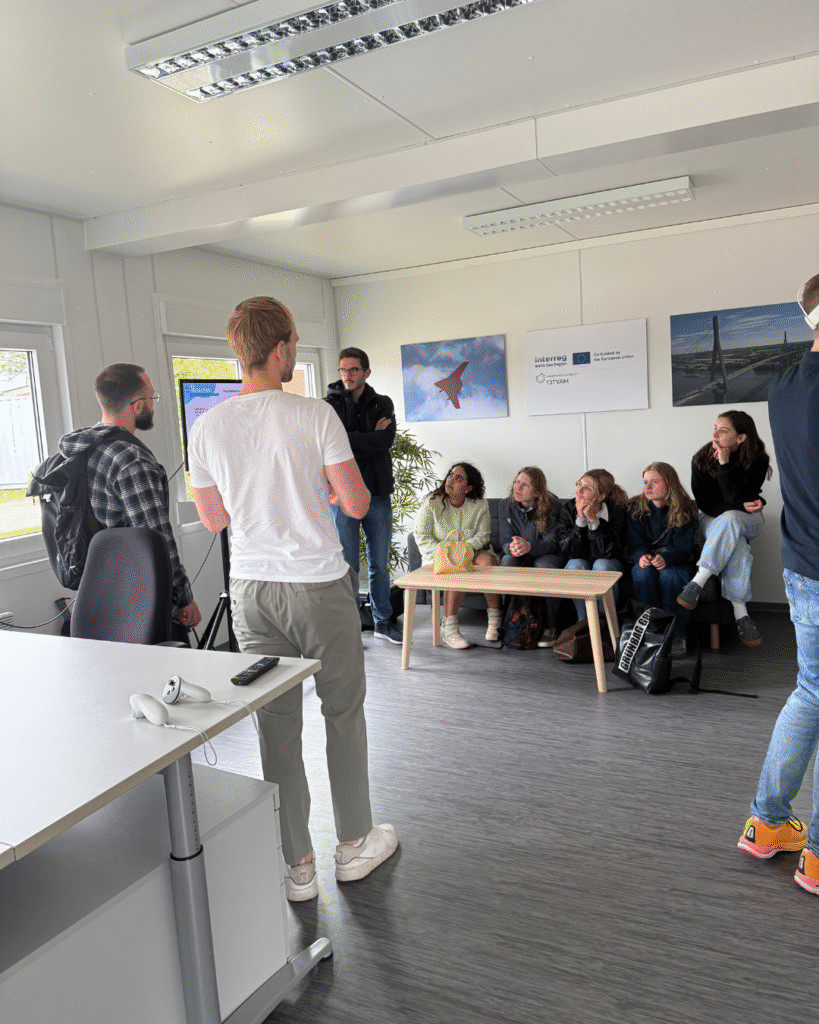 A group of young adults, seated and standing in a bright indoor room, are participating in a discussion or presentation. Some are sitting on a bench or couch, attentively listening, while others stand near the front, possibly presenting. Posters on the wall indicate EU co-funding, and a photo of a jet and a bridge are visible. The space appears to be part of an educational or professional workshop setting.