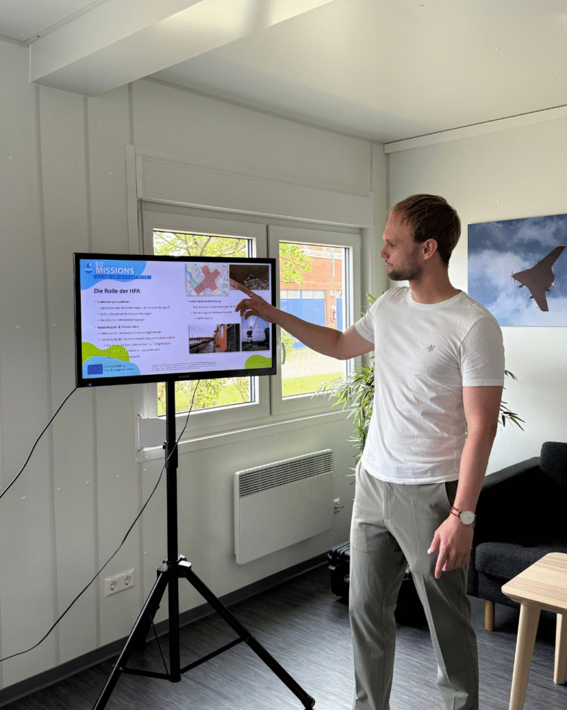 A man in a white t-shirt points at a presentation slide displayed on a screen mounted on a tripod. The slide shows information about EU missions and the role of the HPA, with diagrams and photos.
