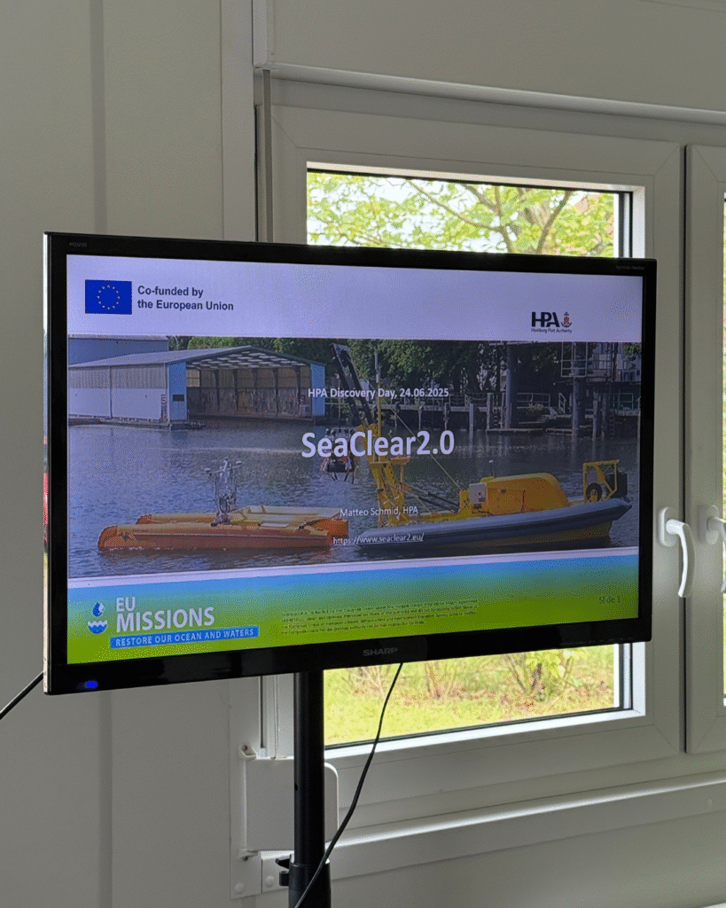 A flatscreen monitor displays a presentation slide titled “SeaClear2.0” with a photo of an autonomousoperated marine vessel. The slide includes logos from the European Union and the Hamburg Port Authority (HPA), and mentions “HPA Discovery Day, 24.06.2025.” The screen is part of an event focused on EU missions to restore oceans and waters.