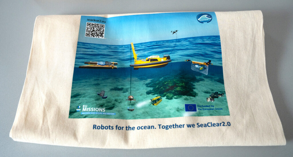 Close-up of a person writing on a green card that says “lack of awareness,” next to a SeaClear2.0 promotional tote bag showing an illustration of marine robotic systems operating underwater.