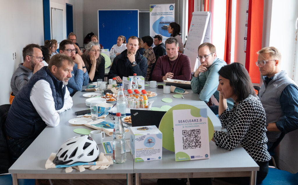 A group of people sitting around a table during a SeaClear2.0 policy workshop, engaged in discussion. The table is covered with drinks, pastries, and informational materials, and a sign reading “SeaClear2.0 Robotic System Components & Technologies” with a QR code is visible in the foreground.