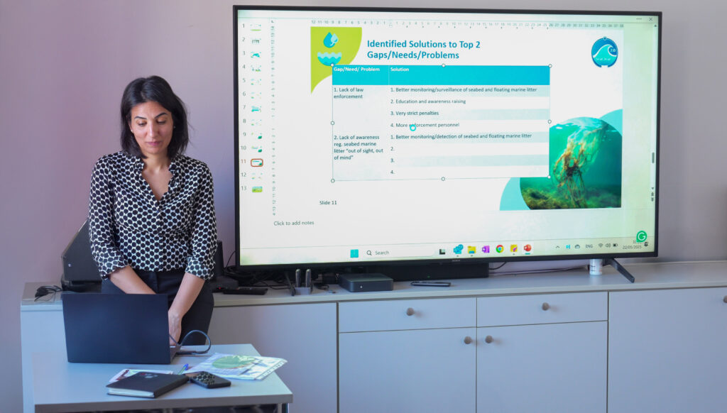 A woman stands in front of a desk with a laptop, while a large screen behind her displays a presentation slide titled "Identified Solutions to Top 2 Gaps/Needs/Problems," listing issues and proposed solutions related to marine litter and law enforcement, along with an underwater image of seagrass.
